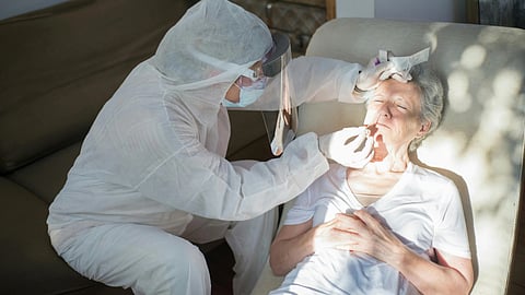 Healthcare worker in protective gear performs a COVID-19 test on an elderly woman lying on a sofa. 
