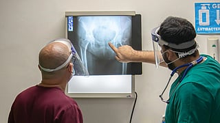 Two healthcare professionals wearing face shields examine a hip X-ray on a lightbox. 