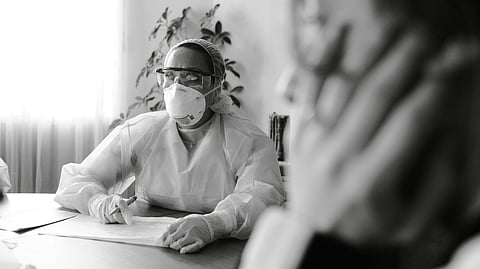 A woman wearing a protective suit and mask sits at a table.