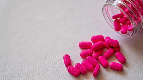 Pink pills spill from a clear bottle onto a smooth, white surface. 