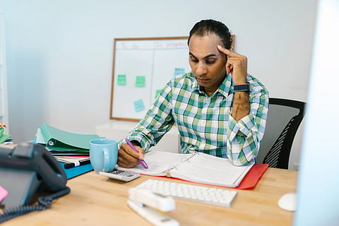 South Asian professional at his office desk, hand to temple, surrounded by paperwork and folders, chronic occupational stress as a driver of metabolic syndrome in urban India