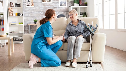 A nurse in blue scrubs kneels beside an elderly woman on a sofa, holding her hand.