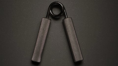 A black hand gripper with textured handles and a metal spring lays on a dark, textured background.