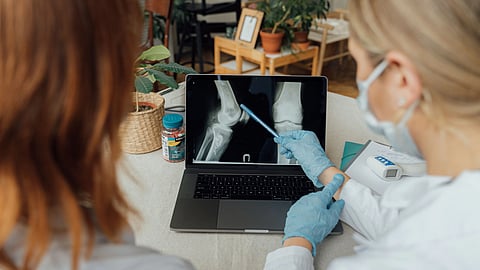 Two people examine a knee X-ray on a laptop. One, wearing gloves, points with a pen.