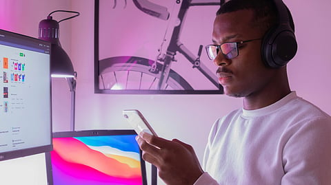 A focused individual with headphones and glasses uses a smartphone in a modern workspace featuring a computer.