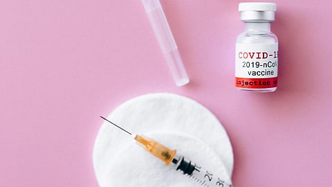 COVID-19 vaccine vials, a syringe, and medical supplies arranged on a pink background, representing childhood vaccination and public health efforts.