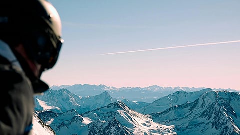 A person in a helmet gazes down at a snowy mountain, contemplating the landscape below.