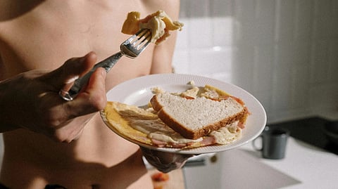 A shirtless man holds a plate with a slice of cake and a fork, smiling as he prepares to enjoy his dessert.