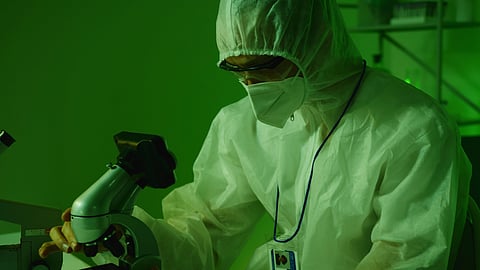 Researcher wearing protective laboratory gear examines samples under a microscope.