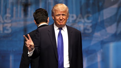 Donald Trump smiles while wearing a dark suit and a bright blue tie, holding up two fingers.