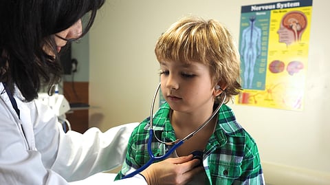 A doctor uses a stethoscope to examine a young boy in a green plaid shirt, in a medical office. 