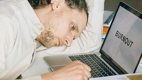 A man rests his head on a laptop while lying on his desk, appearing tired or overwhelmed.