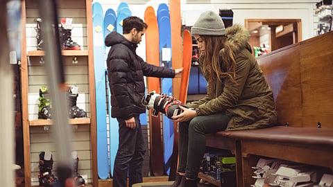 A woman checks ski boots in a ski shop while a man looks at snowboards in the background.
