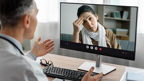A doctor in a white coat gestures while on a video call with a concerned patient. The patient touches her forehead.