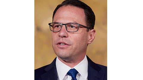 A person in glasses wearing a suit speaks against a plain background.