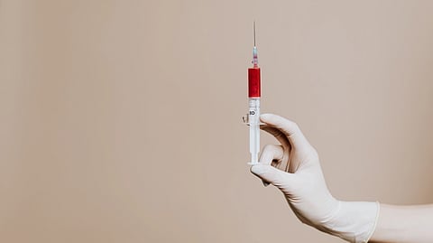 A gloved hand holds a syringe with red liquid against a beige background.