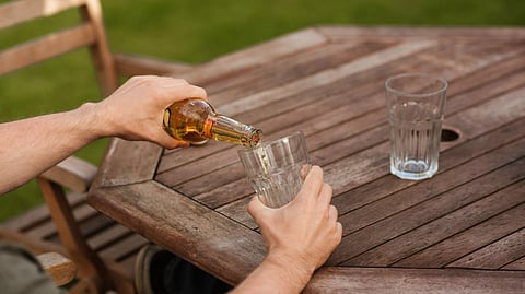 A person pours alcohol from a bottle into a glass on a wooden outdoor table.