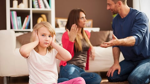 A young girl covers her ears while adults argue in the background.