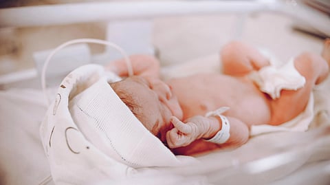 Newborn baby wearing a hat and diaper lies in an incubator, peacefully sleeping.