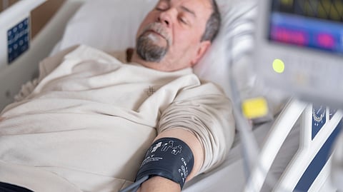 Elderly man resting in hospital bed, eyes closed, wearing a blood pressure cuff on his arm.