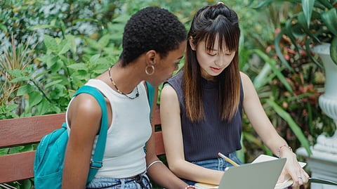 Two young women sit on a bench, focused on a laptop, engaged in conversation.