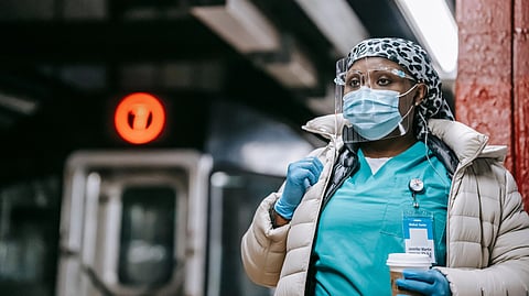 A healthcare worker in scrubs, mask, and face shield stands in a subway station holding a coffee.