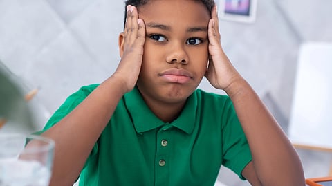 A young boy sitting at a table, resting his hands on his head, appearing deep in thought.