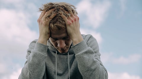 A fair-skinned person with messy blonde hair wearing a grey hoodie is sitting with their head in their hands.