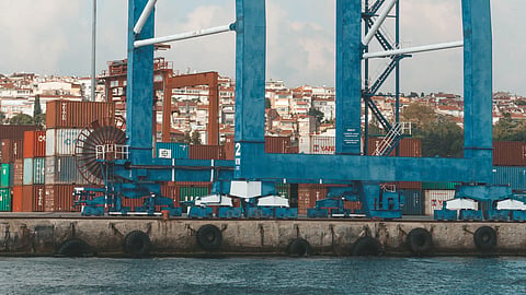 A large, bright blue container crane stands prominently against a light blue sky with scattered clouds.