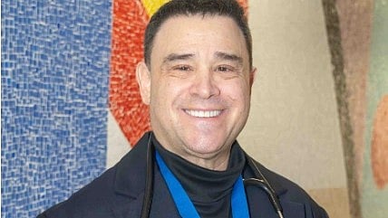 Dr. Mason Blake Pimsler smiles warmly, wearing a navy blazer, black turtleneck, and a stethoscope around his neck.