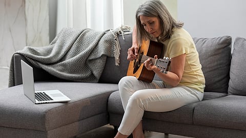 A woman with grey hair is sitting on a grey couch and playing an acoustic guitar.