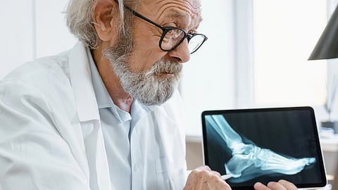 Elderly doctor in a white coat examines a foot X-ray on a tablet.