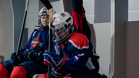 Two hockey players sitting in a locker room wearing full gear.