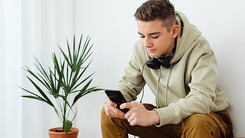 A teenage boy sits by a wall, looking at his smartphone, with headphones around his neck and a small plant beside him.