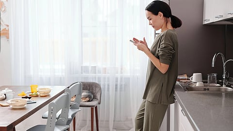 A woman stands in a kitchen, holding her cell phone while looking at the screen.