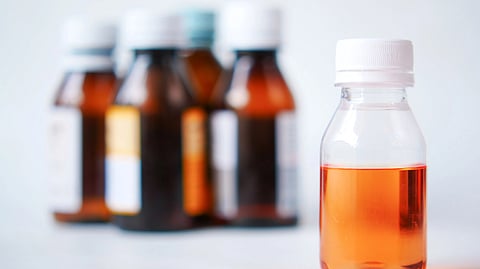 A clear plastic bottle with orange liquid medicine in front, and blurred amber medicine bottles in the background.