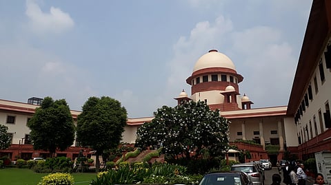 Image of the Supreme Court of India with parked vehicles visible nearby.