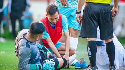 An injured soccer player sits on the field as teammates and a referee check on him.