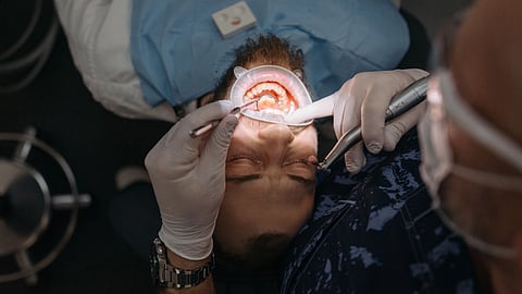 A man sits in a dental chair while a dentist examines his teeth with a dental mirror and tools.