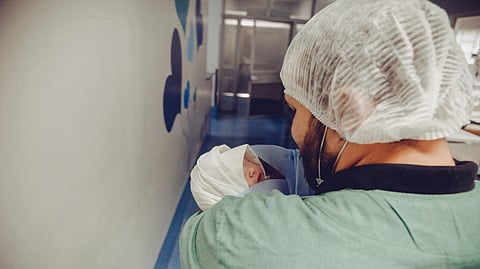 A healthcare worker in scrubs gently holds a newborn baby wrapped in a blanket in a hospital hallway.