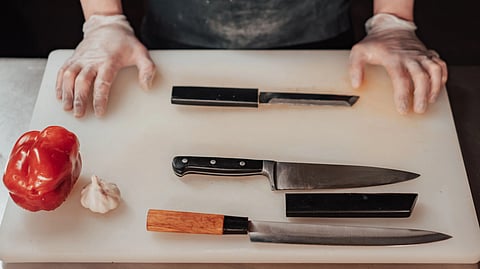 A gloved person stands behind a cutting board with a bell pepper, garlic, and knives.