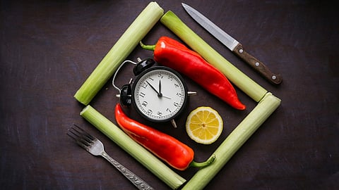 The picture showcases a clock surrounded by food items on a table, along with food cutlery like fork and knife