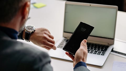 A person in a grey suit sits at a white desk, holding a phone showing 13:02 and checking a wristwatch, with a laptop and papers in front.