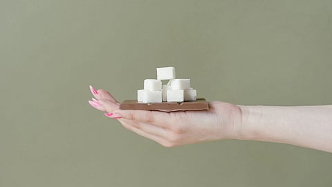 A hand with pink nails holds a piece of chocolate topped with sugar cubes against a green background.