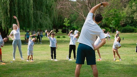 A group of adults and children do stretching exercises in a sunny green park.
