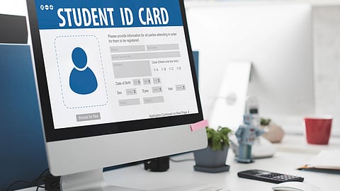 An iMac shows a student ID form on a clean desk with a plant, small robot, and study items nearby.
