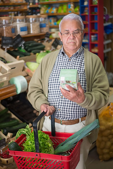 An elderly man with glasses standing in a grocery store aisle, closely reading the back label of a small green box while holding a red shopping basket containing fresh green lettuce and leeks.