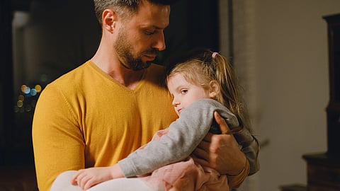 A man in a yellow sweater gently holds a sleepy young girl in a warm, caring moment.