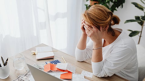 A stressed woman sits at a desk with bills, papers, and a calculator around her.