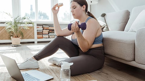 A woman sits on the floor, using a laptop while holding a dumbbell in her right hand.